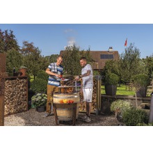 Zwei Personen grillieren im Garten mit einem Fassgrill, umgeben von Holzregalen und Pflanzen.