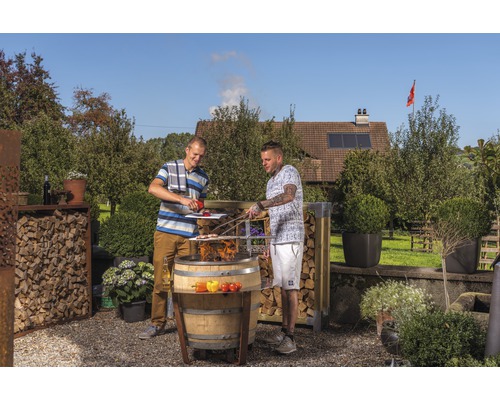 Zwei Personen grillieren im Garten mit einem Fassgrill, umgeben von Holzregalen und Pflanzen.