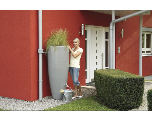 Szene mit Regenwassertank, Bepflanzung und Giesskanne vor einem Haus.