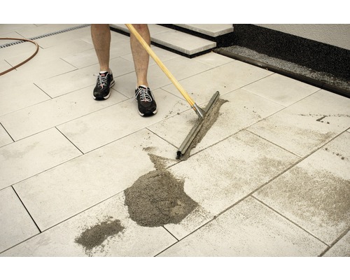 Eine Person verteilt Fugensand mit einem Gummischieber auf einer gepflasterten Fläche.