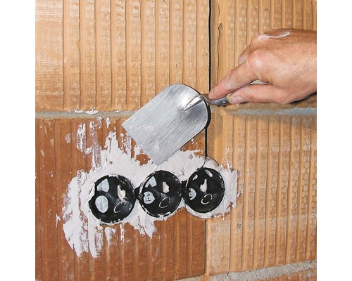 Handwerker verspachtelt eine Wand aus Ziegelsteinen mit drei schwarzen Elektrodosen