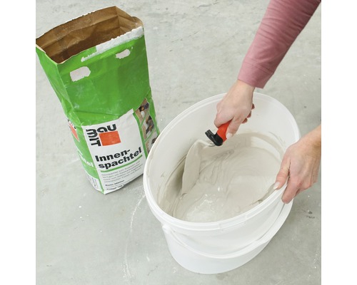 Enduit intérieur mélangé dans un seau avec une spatule, avec un sac d''enduit intérieur Baumit en arrière-plan