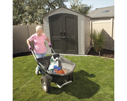 Eine Frau schiebt eine Schubkarre mit Gartengeräten und -materialien vor einem Gartenschuppen.