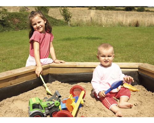 Deux enfants jouent avec des moules à sable et un tracteur jouet dans un bac à sable en bois dans le jardin.