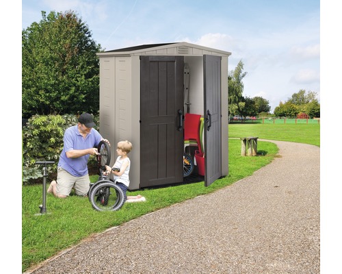 Gartenszene mit Gerätehaus, in dem Gartengeräte und ein Kinderfahrrad aufbewahrt werden, während ein Vater mit seinem Sohn ein Fahrrad repariert