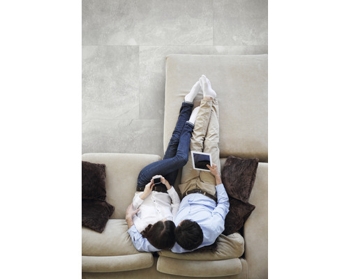 Ein Paar entspannt auf einem Sofa mit Tablets und Handys, während graue Bodenfliesen im Hintergrund zu sehen sind