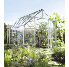 Freistehendes Gewächshaus aus Aluminium mit Glasscheiben und Dachfenster in einem sonnigen Garten mit Blumenbeeten und Gartengeräten.