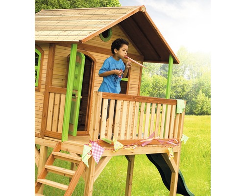 Maisonnette en bois avec toboggan et un enfant sur la véranda soufflant dans un chapeau de fête