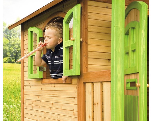 Un enfant joue avec une trompette dans une cabane en bois avec des fenêtres vertes.