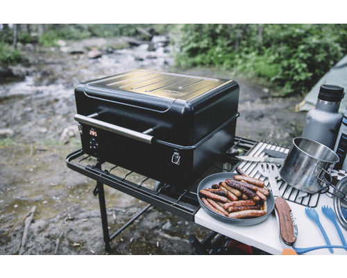 Tragbarer Grill mit digitaler Anzeige und Würstchen auf einem Teller im Freien.