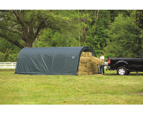 Graue Lagerhalle für Heuballen auf einer Wiese, eine Person beim Ausladen eines Heuballens in der Nähe eines Pickups.