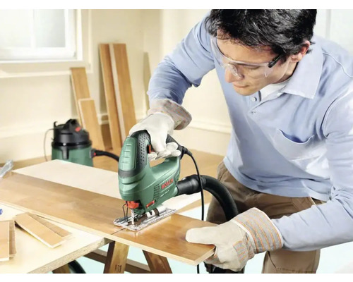 Un homme scie une planche de bois avec une scie sauteuse, des lunettes de protection et des gants