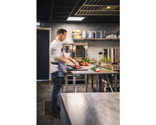 Un cuisinier prépare des légumes et de la viande sur une table en acier inoxydable dans une cuisine professionnelle.