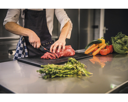 Une personne coupe de la viande sur une planche à découper, entourée de légumes et d''herbes aromatiques.