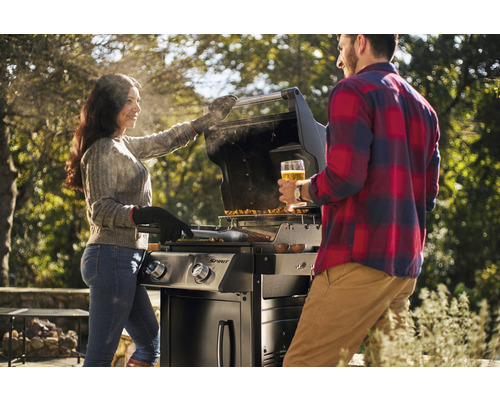 Ein Paar grilliert mit einem Gasgrill im Garten. Die Frau wendet das Grillgut, der Mann hält ein Glas Bier.
