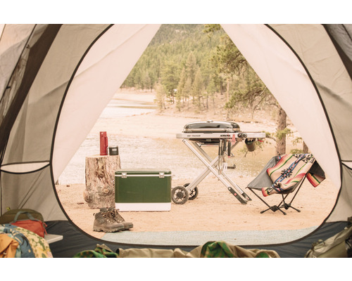 Campingplatz mit Grill, Kühlbox, Thermoskanne und Stiefeln, gesehen aus einem Zelt