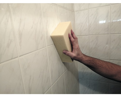 Eine Hand hält einen Schwamm an eine geflieste Wand.