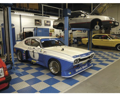 Scène d''un garage avec plusieurs voitures anciennes, dont une Ford Capri avec des accents bleus et le numéro trois.