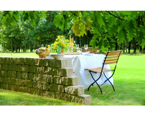 Gedeckter Tisch im Garten mit Steinmauer, Stühlen, Blumen und Speisen.