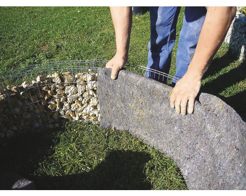Une personne fixe un voile de séparation à une cage de pierres dans le jardin.