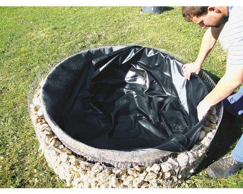 Un homme installe une bâche de bassin dans une cage de gabions ronde remplie de pierres