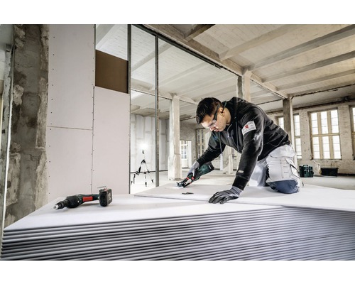 Un artisan coupe une plaque de plâtre avec un outil spécial dans une construction brute, d''autres outils étant prêts à l''emploi.