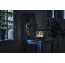 Lanterne à boule à neige décorative avec motif de village d''hiver sur une table, entourée de plantes et d''une couronne.