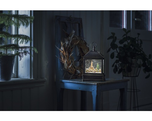 Lanterne à boule à neige décorative avec motif de village d''hiver sur une table, entourée de plantes et d''une couronne.