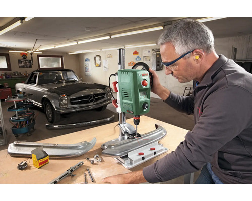 Une personne utilise une perceuse à colonne dans un atelier avec une voiture ancienne en arrière-plan.
