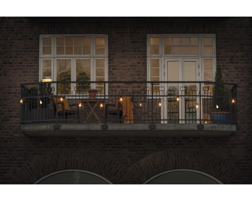 Balcon décoré avec guirlande lumineuse, chaises, table et plantes devant un bâtiment en briques.
