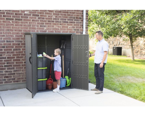 Gartenschrank mit Werkzeugen und Gartenutensilien, ein Kind räumt den Schrank ein, ein Erwachsener hält die Tür auf dem Rasen