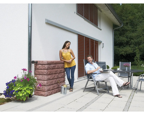 Szene mit Regenwassertank in Steinoptik, Blumen, Paar auf der Terrasse und Hausfassade