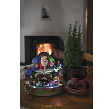 Weihnachtliche Dekoration mit Weihnachtsmann, Kindern, Zug, kleinem Baum und Dekokugeln auf einem Tisch vor einem Kamin.