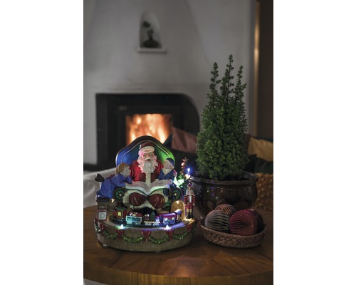 Weihnachtliche Dekoration mit Weihnachtsmann, Kindern, Zug, kleinem Baum und Dekokugeln auf einem Tisch vor einem Kamin.