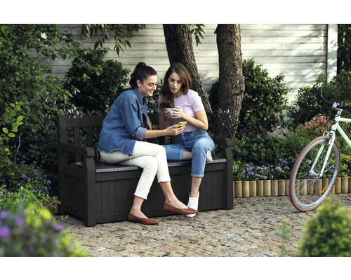 Deux femmes sont assises sur un banc de jardin avec espace de rangement et regardent un smartphone.