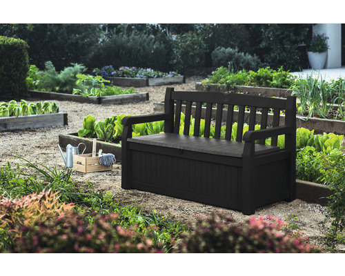 Scène de jardin avec un banc de jardin noir avec fonction de rangement et un panier en bois avec des outils de jardin.