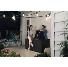Un couple est assis sur un coffre de jardin noir sur une terrasse sous une guirlande lumineuse.