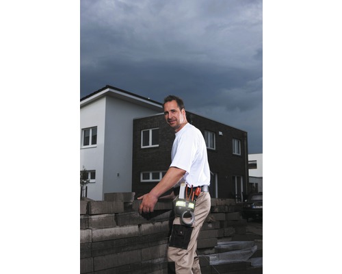 Ein Bauarbeiter mit Werkzeuggürtel hantiert mit Pflastersteinen vor einem Haus.