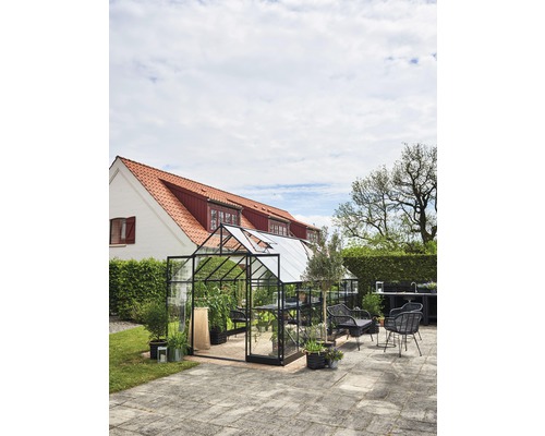 Gewächshaus mit Pflanzen und Gartenmöbeln auf einer Terrasse vor einem Haus.