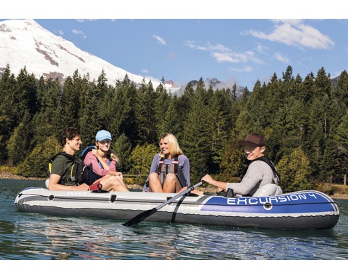 Quatre personnes dans un bateau pneumatique Intex Excursion 4 sur un lac devant un paysage de montagnes boisées.