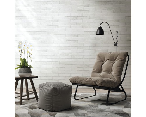 Wohnzimmer-Szene mit Sessel, Fußhocker, Beistelltisch, Stehlampe und Dekoration auf einem gemusterten Teppich vor einer Steinwand.