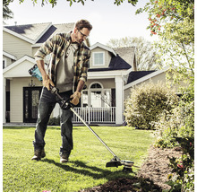 Mann trimmt Rasen mit einem Akku-Rasentrimmer im Garten vor einem Wohnhaus.