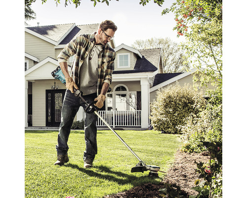 Mann trimmt Rasen mit einem Akku-Rasentrimmer im Garten vor einem Wohnhaus.