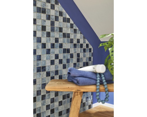 Mur de salle de bains avec des carreaux de mosaïque dans différentes nuances de bleu, serviette et décoration sur un tabouret en bois.