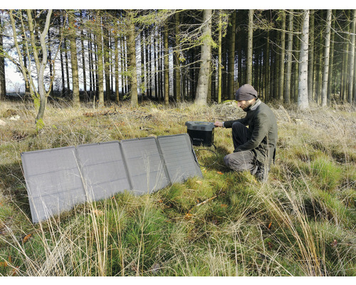 Un homme est accroupi dans la forêt et utilise un module solaire pour charger un générateur.