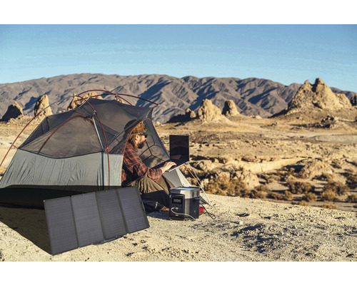 Un homme travaille dans une tente avec un ordinateur portable, un générateur et un module solaire dans un paysage de montagne.