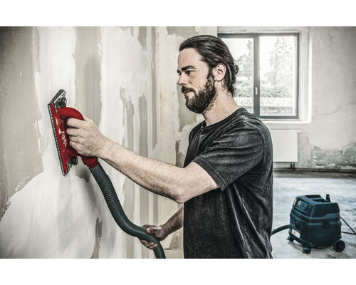 Handwerker schleift Wand mit einem Handschleifer, der an einen Staubsauger angeschlossen ist.
