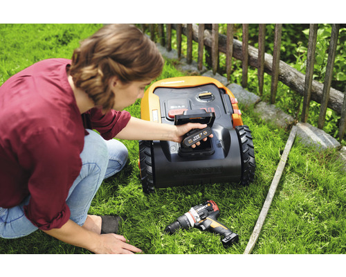 Une femme insère une batterie dans un robot de tonte, un tournevis sans fil est posé dans l''herbe