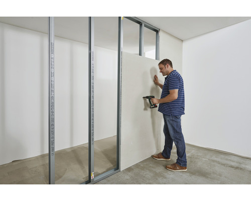 Un homme fixe une plaque de plâtre avec une perceuse sur des profilés métalliques dans un bâtiment brut.