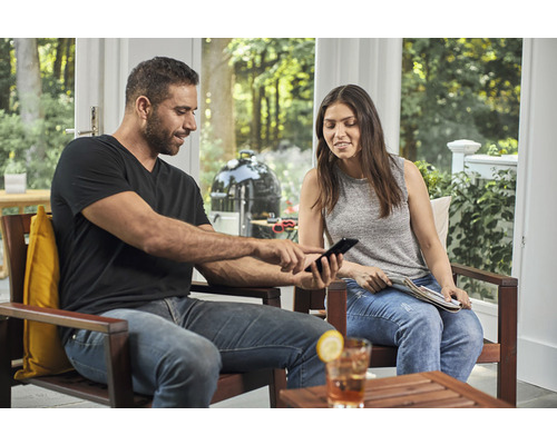 Un homme et une femme sont assis sur la terrasse et regardent un téléphone portable, avec un barbecue sphérique en arrière-plan.